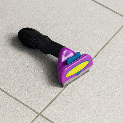 Brosse pour chat SoyeuxPro à tête violette, bouton bleu et manche noir, posée sur un carrelage beige