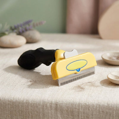Brosse jaune pour chat à poil long, avec manche noir et dents métalliques, posée sur une surface en tissu beige