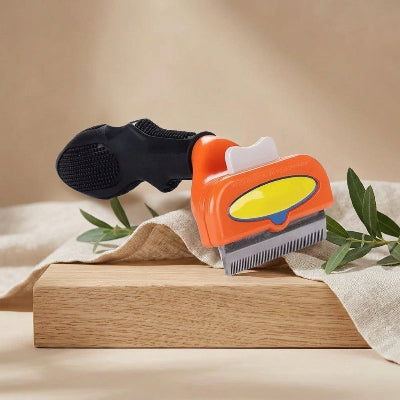 Brosse orange pour chat à poil long, avec manche noir et dents métalliques, présentée sur un support en bois avec un tissu beige et des branches vertes